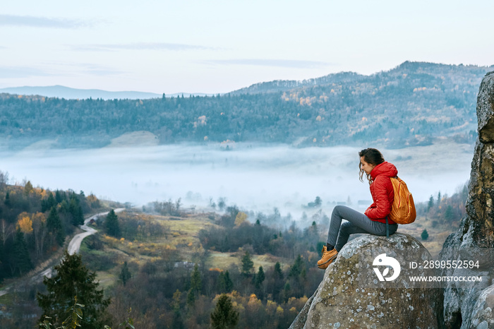 woman hiker with backpack, wearing in red jacket, sitting on edge of cliff against a forest and foggy valley background