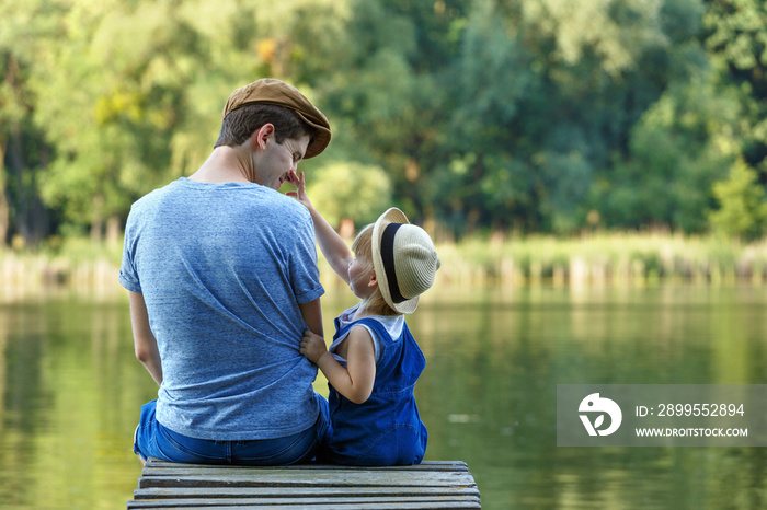 Father and a tiny daughters in dungarees are sitting on a river bridge and looking at each other. They talk, laugh and have fun.