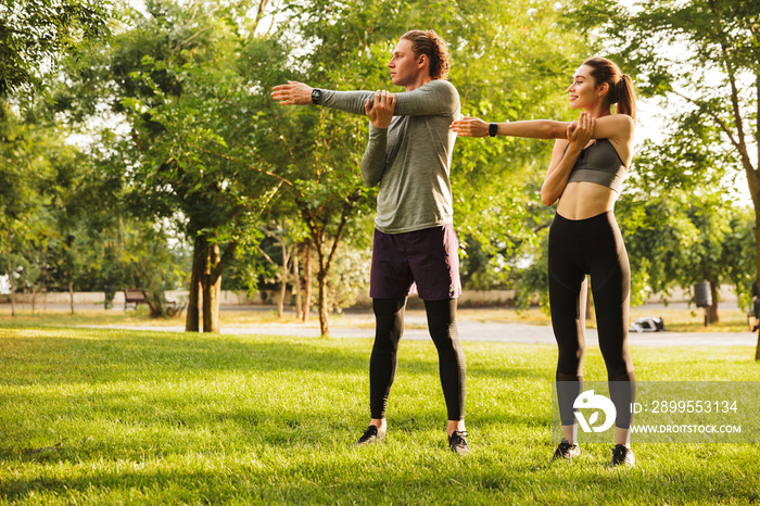 Photo of fitness sportsman and sportswoman 20s in tracksuits, warming up together and stretching arms in green park during sunny summer day