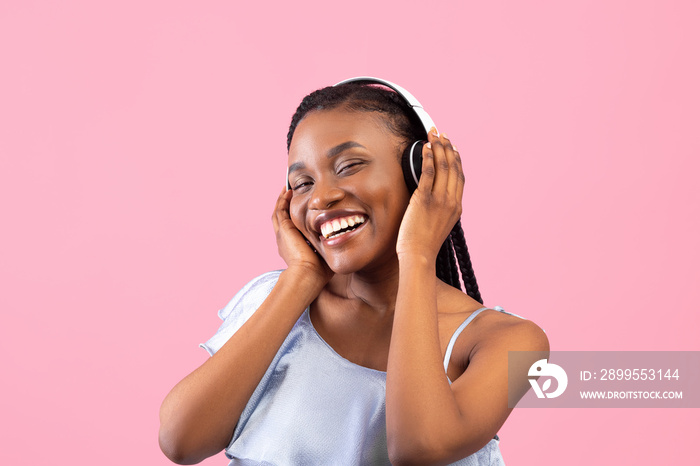 Relaxed black woman wearing headphones, listening to modern music on pink studio background