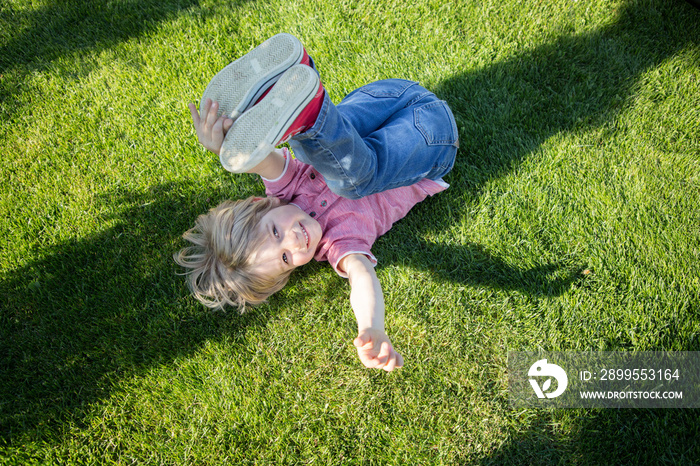 Happy little boy lies upside down on the green grass in the park. Children’s happiness, joy, self-indulgence. Good mood, relax