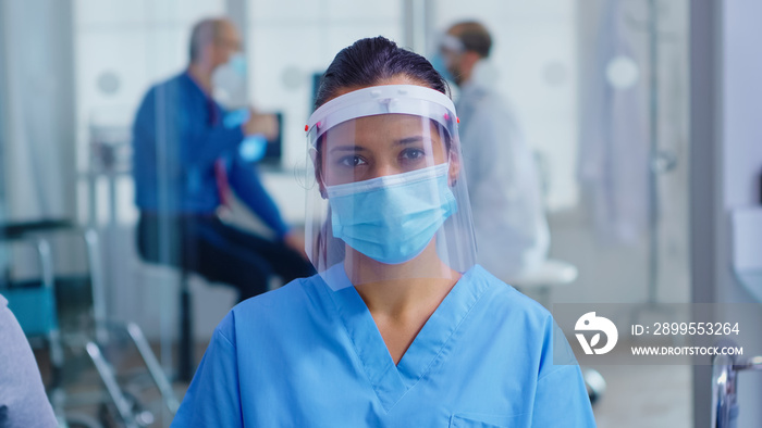 Medical assistant with visor and face mask against coronavirus looking at camera in hospital waiting area. Doctor consulting senior man in examination room.