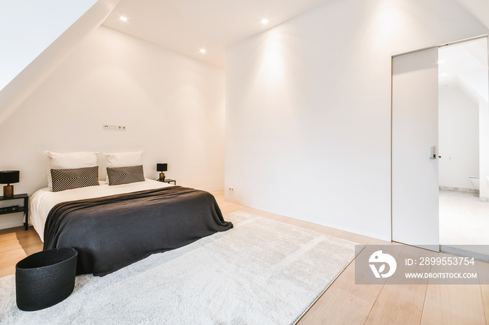 Spacious white colored bedroom with big bed and tables under attic roof with carpet and cozy armchair