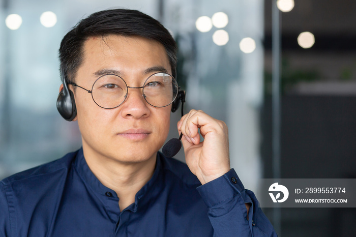 Close up photo. Portrait of a serious Asian man in a headset. He looks into the camera, holds the microphone in his hand. Businessman, business trainer, saleman, customer support hotline.