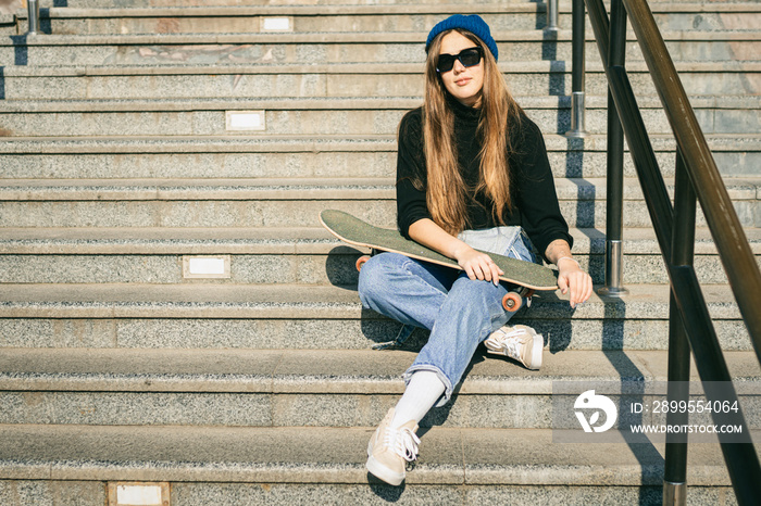 Urban woman with skate. Hipster girl with skateboard in city. Extreme sport and emotions concept. Alternative lifestyle. Stylish hipster girl holding skateboard and posing. Street style outfit