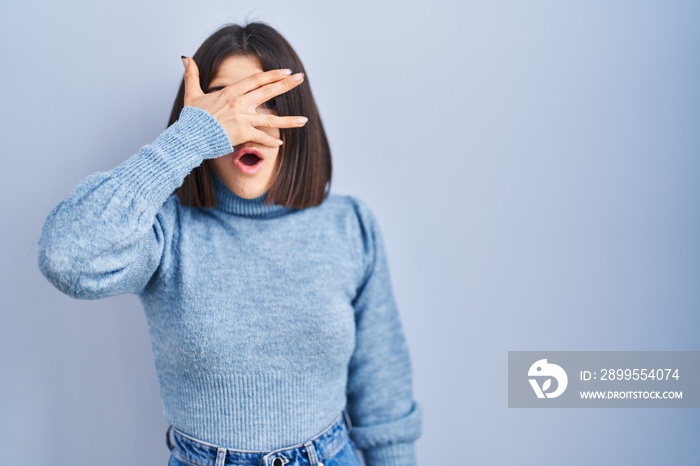 Young hispanic woman standing over blue background peeking in shock covering face and eyes with hand, looking through fingers with embarrassed expression.