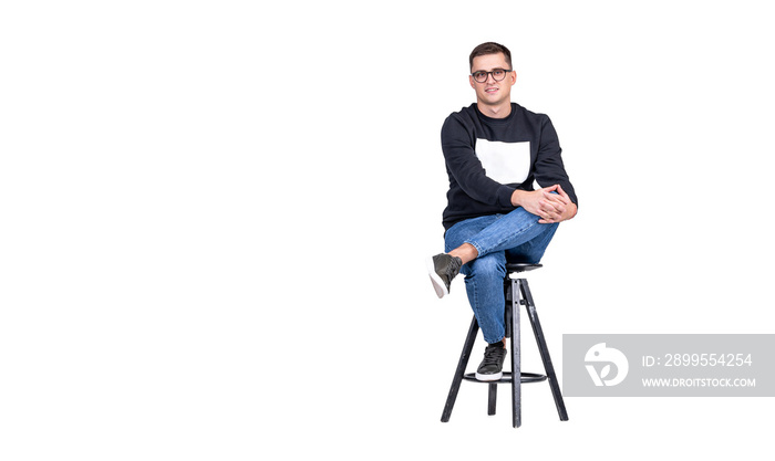 A young attractive guy in blue and black clothes sits on a bar stool on a white background and looks at camera. Copy space.