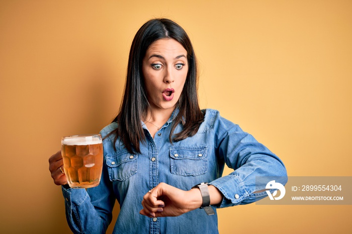 Young woman with blue eyes drinking jar of beer standing over isolated yellow background Looking at the watch time worried, afraid of getting late