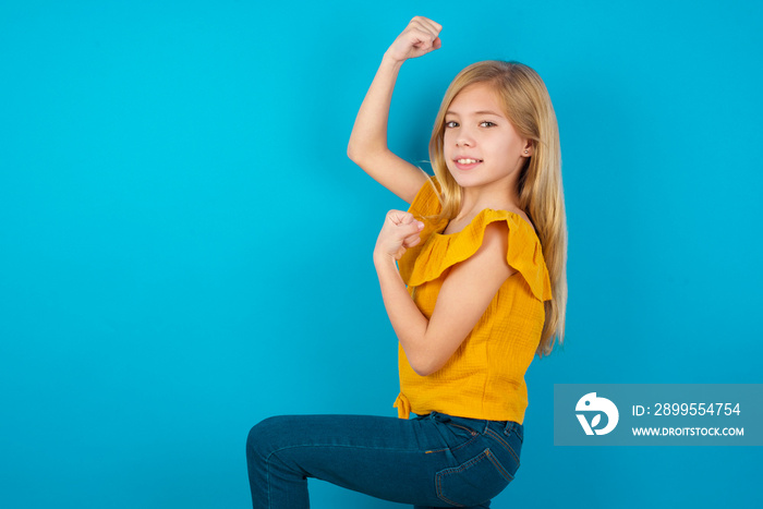 Profile photo of excited Caucasian kid girl wearing yellow T-shirt against blue wall raising fists celebrating black Friday shopping