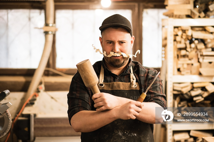 Smiling, funny male carpenter work in cap and coveralls with shaving mustache, holding crossed chisel and wooden hammer