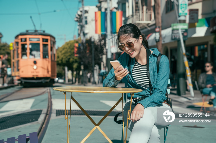 happy beautiful girl tourist sitting at outdoor cafe table and using smart phone chatting online with friends. young woman relax on street with cable car in background. castro district in usa.