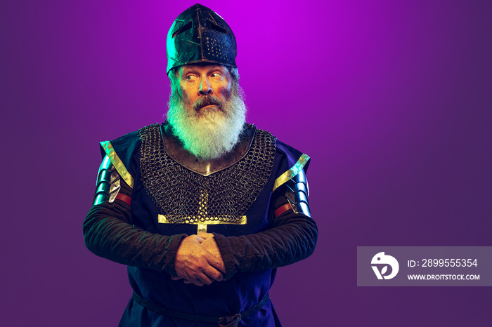 Comic portrait of senior grey-bearded man in image of medieval warrior or knight in armor with sword isolated over purple background in neon. Comparison of eras