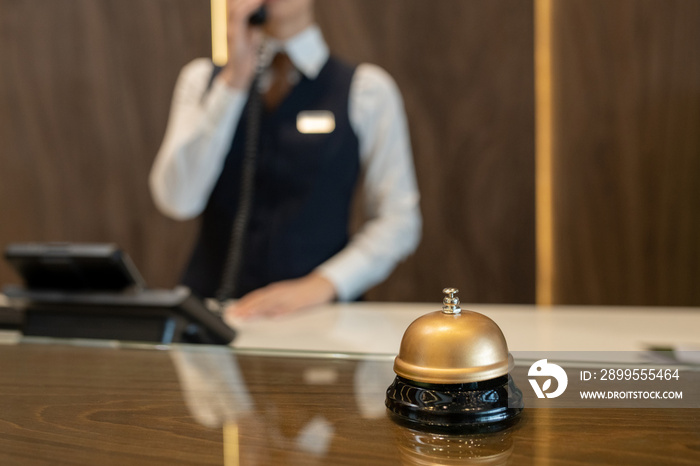 Bell on wooden reception counter against female receptionist consulting clients on the phone while working in contemporary luxurious hotel