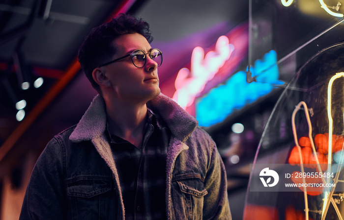 A young man fashionably dressed standing in the street at night. Illuminated signboards, neon, lights.