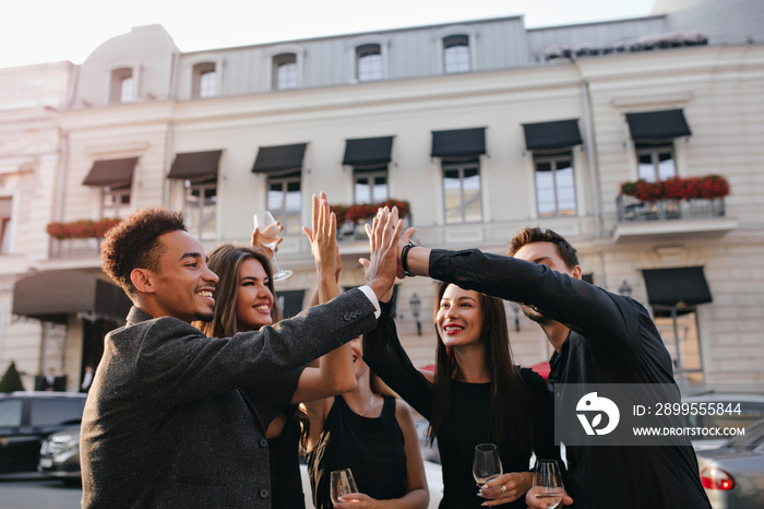 International friends slap hands enjoying team work. Outdoor portrait of colleagues celebrating successful project with champagne posing in front of office building.