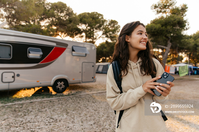 Positive young caucasian girl smiling looking away, holding phone standing outdoors. Brunette wears hoodie and backpack. Technology concept, lifestyle