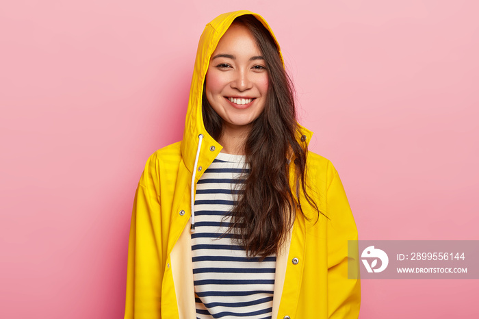 Smiling beautiful woman enjoys wearing warm striped jumper, yellow raincoat with hood, has good mood, goes out with friends during rainy day, poses indoor. People, weather and season concept