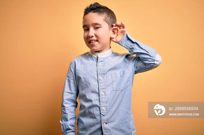 Young little boy kid wearing elegant shirt standing over yellow isolated background smiling with hand over ear listening an hearing to rumor or gossip. Deafness concept.