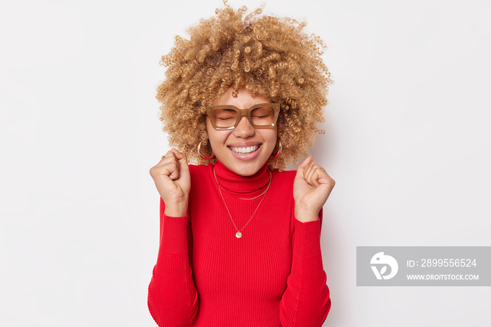 Winning concept. Positive curly haired young woman clenches fists in triumph smiles toothily keeps eyes closed wears spectacles and casual red poloneck isolated over white background. Yeah go win