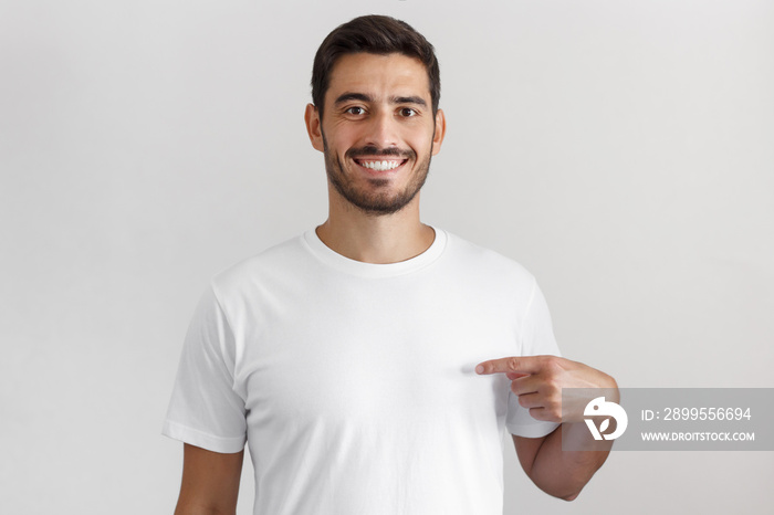 Daylight portrait of smiling nice man pointing at his blank white t-shirt with index finger, copy space for your advertising, isolated on grey background