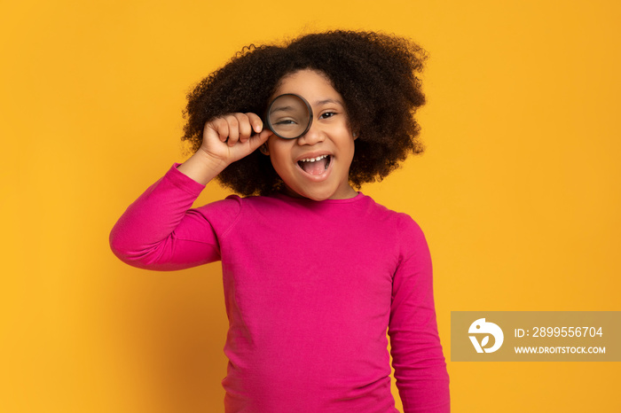 Beautiful little african girl looking through magnifying glass on yellow background