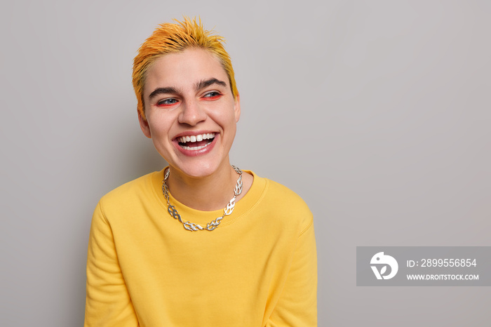 Portrait of positive hipster with yellow hairstyle bright vivid makeup dressed in casual jumper metal chain has own believes extraordinary look poses against grey background blank copy space