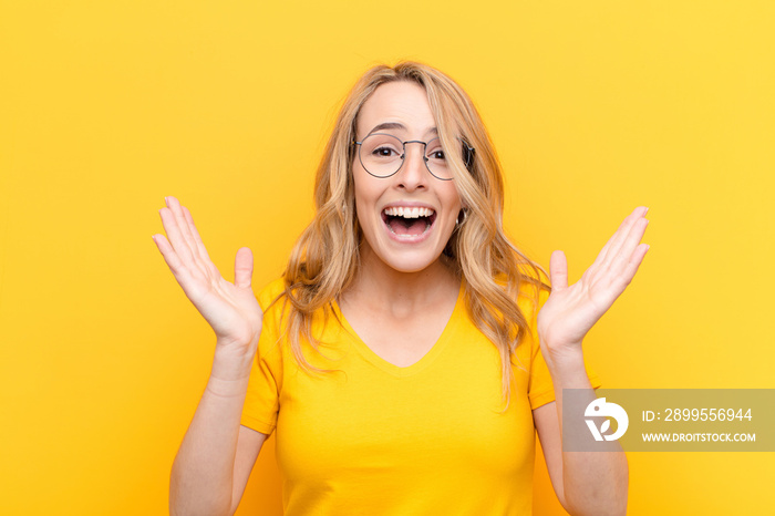 young pretty blonde woman looking happy and excited, shocked with an unexpected surprise with both hands open next to face against flat color wall