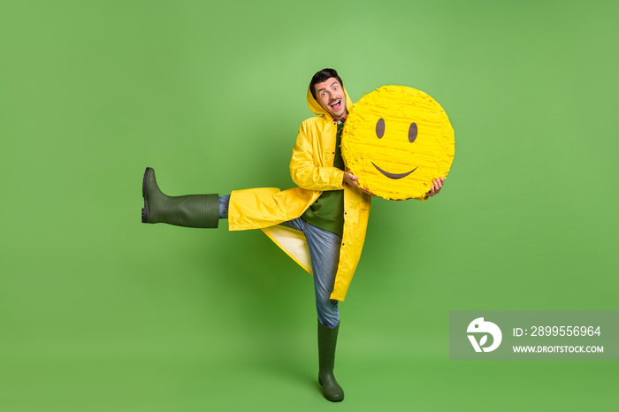 Full length photo of young cheerful man happy positive smile hold paper pinata emoji wear raincoat isolated over green color background