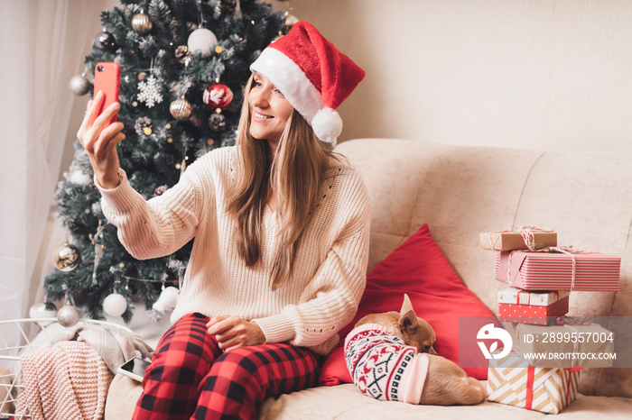 Young Woman and dog in sweater takes selfie portrait on the smartphone at winter holidays, enjoy Christmas time at home. Wrapping gifts and video chat at mobile phone.