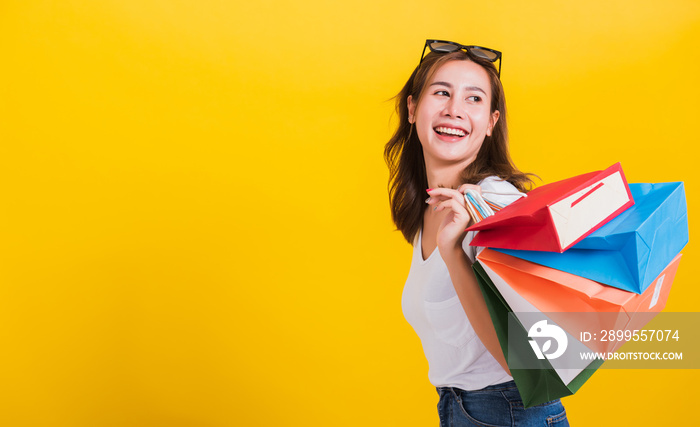 Asian Thai portrait happy beautiful cute young woman smiling stand with sunglasses excited holding shopping bags multi color looking back, studio shot isolated yellow background with copy space