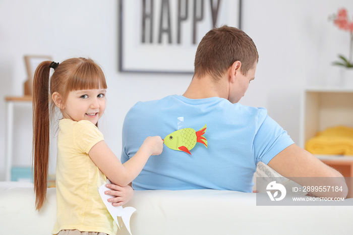 Little girl sticking paper fish to her father’s back indoors. April fool’s day prank