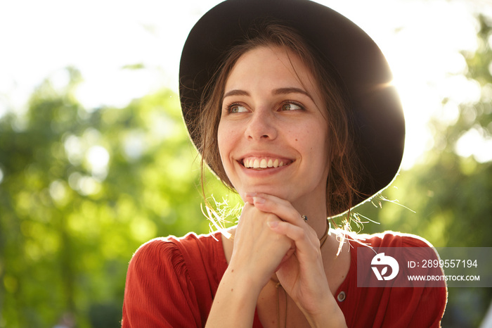 Close-up portrait of pleased woman with pure skin, dark eyes and sincere smile wearing summer hat and red shirt isolated over nature background. Dreamy woman enjoying sunlight and fresh summer air