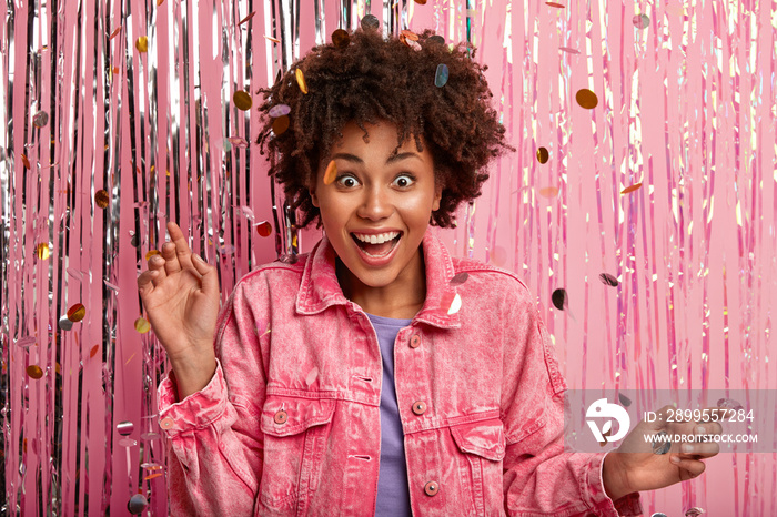 Indoor shot of happy surprised dark skinned lady with Afro haircut, dressed in pink fashionable jacket, poses over confetti and tinsel background, being on bash, feels surprised to be invited on dance