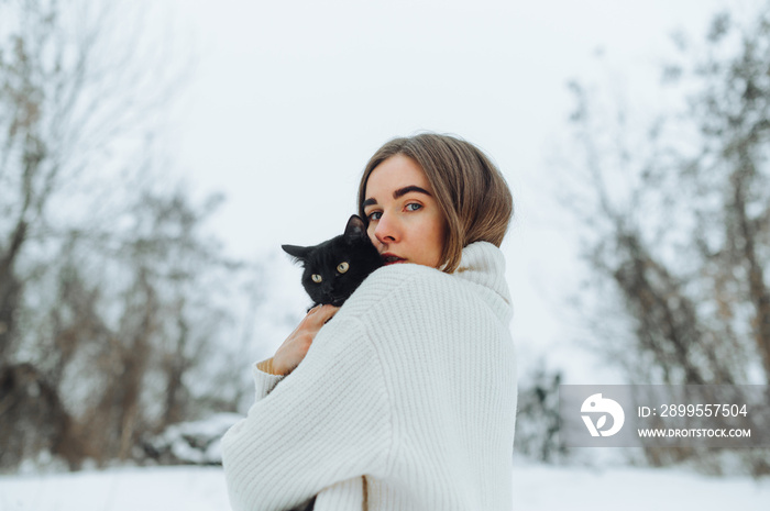 Beautiful woman in a knitted sweater posing with a black cat on a walk on a snowy street, looking at the camera with a serious face