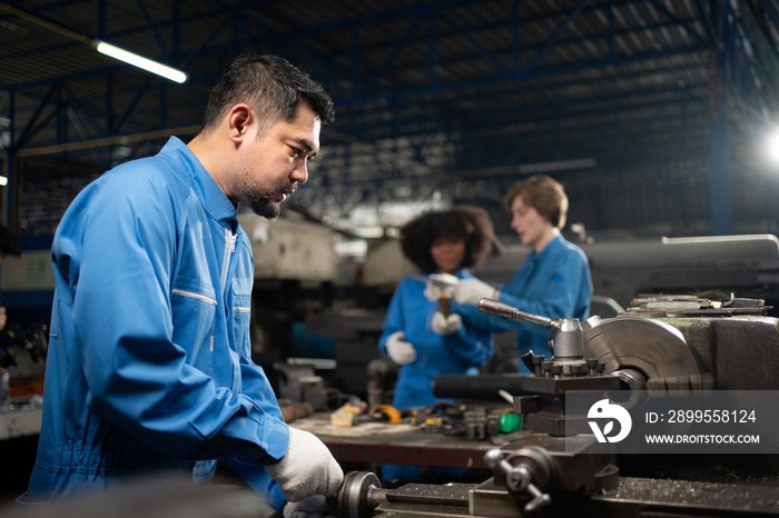 Selective focus of Asian male lathe worker in a blue uniform and gloves, standing working with cross feed handwheel of a lathe machine with blurred colleagues working in background in a metal factory.