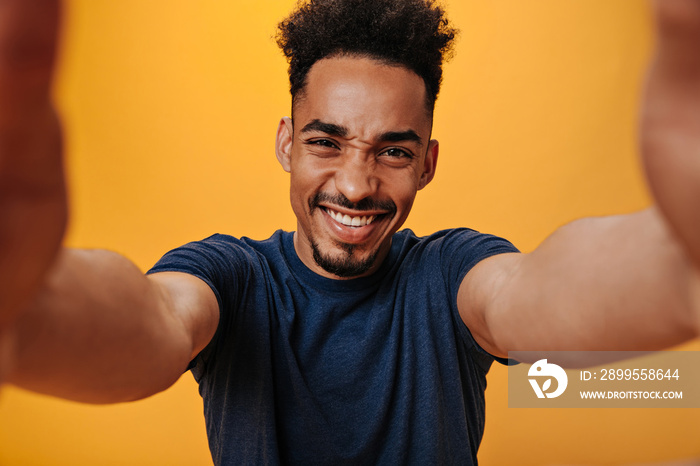 Joyful guy in blue T-shirt makes selfie on isolated background. Snapshot of cool man with tattoo posing on orange backdrop