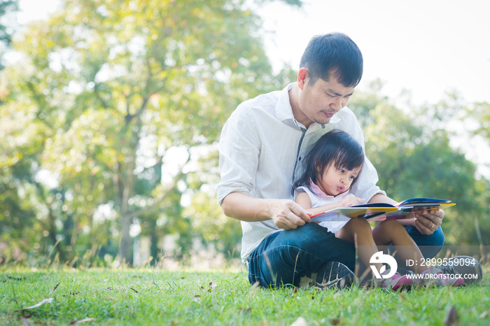 Asian father and daughter are reading the book together in the park with fully happiness moment, concept of learning activity for kid in family lifestyle.