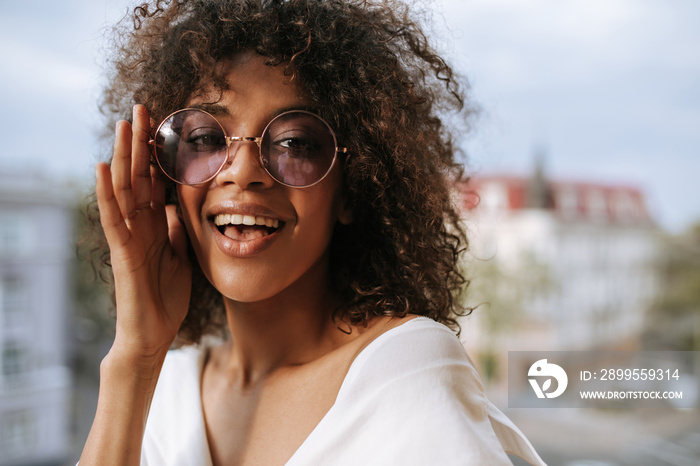 Attractive girl in round eyeglasses smiling on terrace. Wavy haired woman in white clothes looking into camera on balcony..