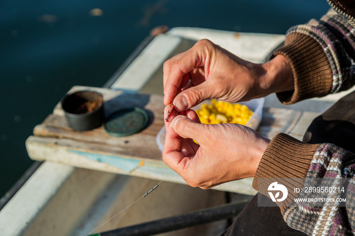 Close up of hands setting worm as bait on fishing hook. Man is holding fishing rod and line with sinkers. He is working on boat while floating over river. Bowl of corn is in background.