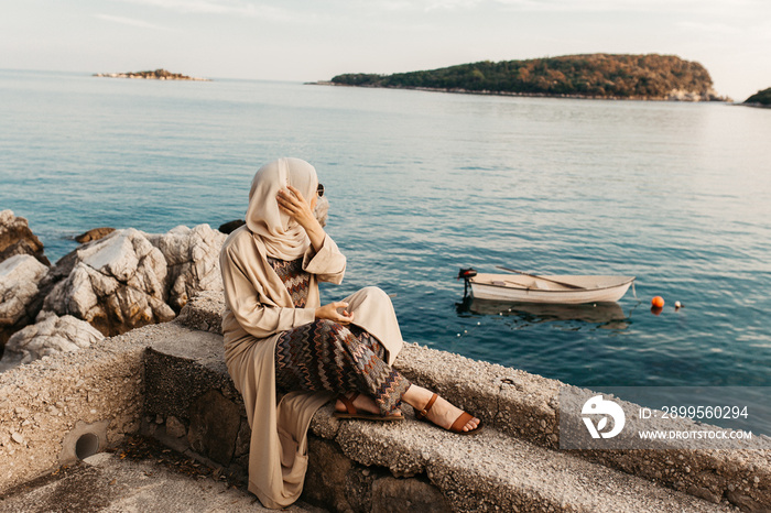 portrait of young European Muslim women with hijab sitting on the stone beach with sea in the background. She is happy and relaxed.