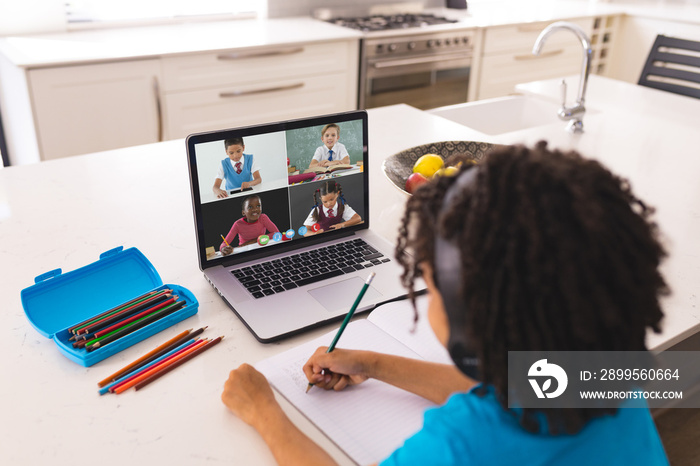 African american boy with curly hair writing notes while learning from online class over laptop