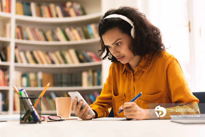 Remote learning. Lady in wireless headphones sitting at desk in library, using smartphone and writing in her notebook