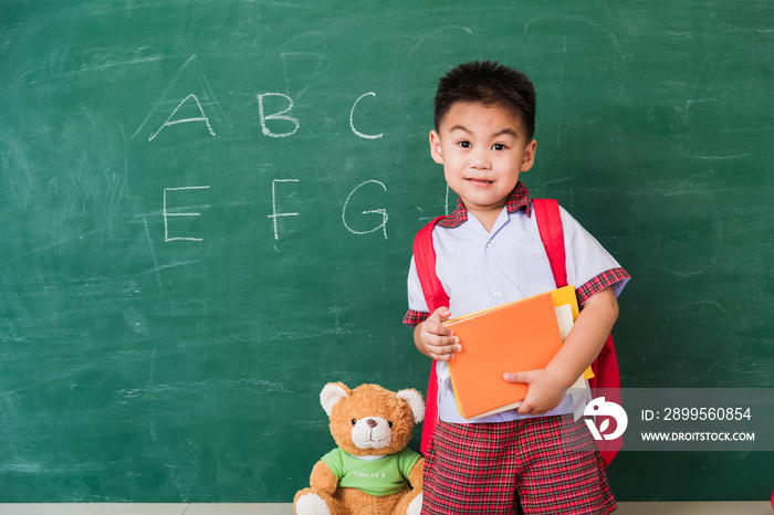 Back to School. Happy Asian funny cute little child boy from kindergarten in student uniform with school bag hold books smile on green school blackboard, First time to school education concept