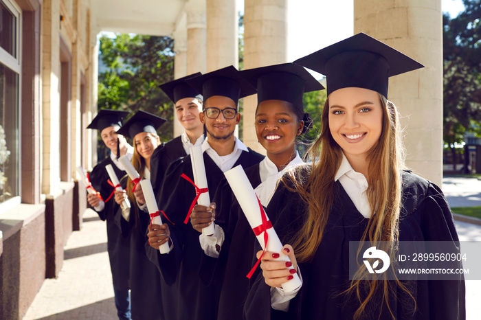 Multinational group of attractive students-graduate in mortar boards cap and ceremonial robes holding diploma in their hands, look very positive and happy, smiling snow-white smile at the camera.