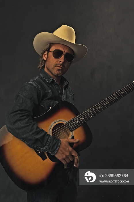 Man in denim shirt, white cowboy hat and aviator sunglasses with acoustic western guitar in front of a grey wall.