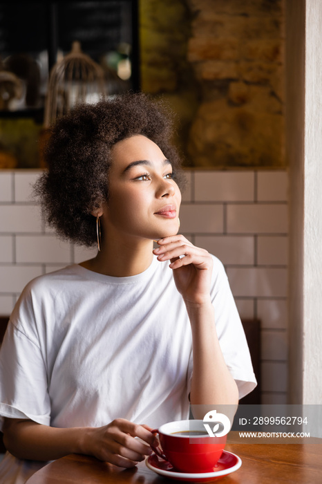 pensive african american woman in white t-shirt holding cup while looking away