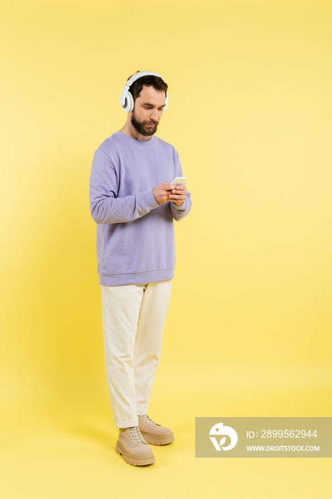 full length of trendy bearded man in wireless headphones using mobile phone on yellow.