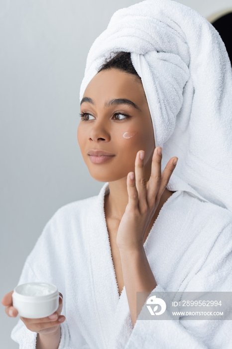 dreamy african american woman in bathrobe holding container with face cream in bathroom