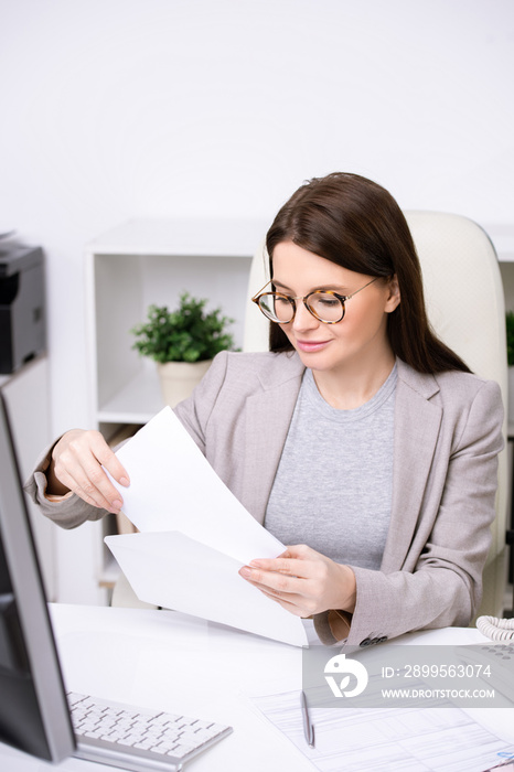 Young businesswoman in glasses sitting at table and putting document into envelope while sending business letter