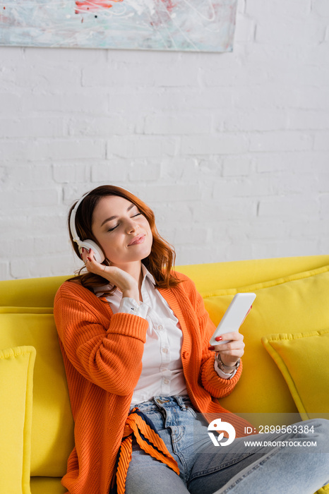 smiling woman in headphones holding smartphone while listening music with closed eyes